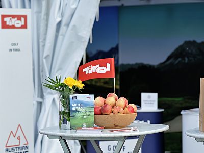 Table with peaches, yellow flowers, Tirol mini flag, golf promo materials, and scenic backdrop, suggesting a promotional event setting.