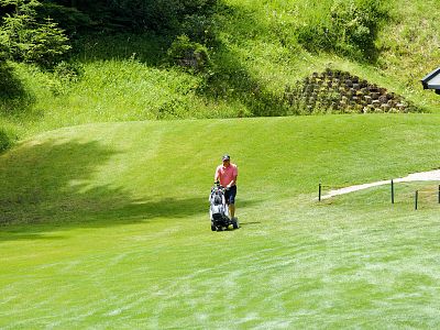 A golfer in a red shirt pushes a cart on a green course, surrounded by trees, on a sunny day. There's a small building to the right.