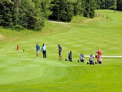 Golfers on a green golf course under a clear sky, with trees surrounding the fairway, some preparing to putt near a red flag.