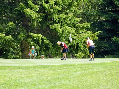 Three golfers on a lush green golf course, surrounded by tall trees. One is putting, while the others watch, near the hole with a flag.