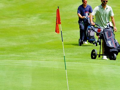 Two golfers on a green course with a flag. Both push their golf bags on trolleys across the putting area, getting ready for their next shots.