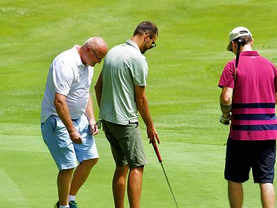 Three men on a golf green, one prepares to putt, while others watch. They wear casual golf attire. Bright, sunny day with lush green grass.