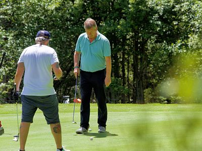 Two people on a golf course, one putting while the other observes. They are surrounded by bright green trees and grass under a sunny sky.