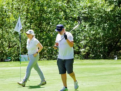 Two individuals on a golf course; one is holding a club, the other checks a phone. They're surrounded by greenery on a sunny day.