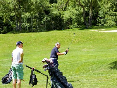 Two men on a golf course, one swinging a club, the other watching. Golf bags are present, with trees in the background under a clear sky.