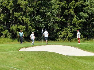 Four golfers walking on a green golf course near a sand bunker, with trees in the background.