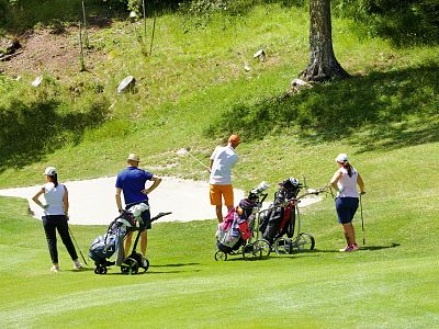A group of five people on a golf course, with two pushing carts and one preparing to swing, surrounded by green grass and a sand trap in the background.