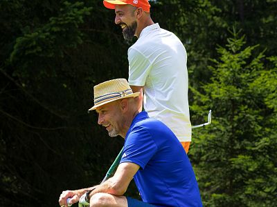 Two men on a golf course, one in a blue shirt with a hat, the other in white, smiling and enjoying the outdoors, surrounded by greenery and trees.