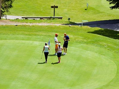 Four people stand on a golf course green, engaged in conversation or focusing on a golf task, surrounded by lush grass and distant trees.