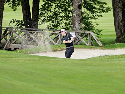 Golfer swings from sand trap, surrounded by green grass and trees with a wooden bridge in the background.