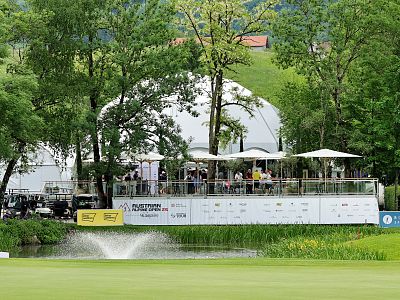 Outdoor event venue by a pond with a fountain, surrounded by trees. A large tent and a viewing platform with people are in the background.