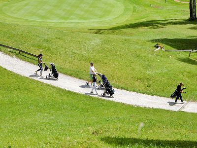 Four people walk on a narrow path through a green golf course, each pushing a golf cart. The course is surrounded by lush grass and a putting green is visible.
