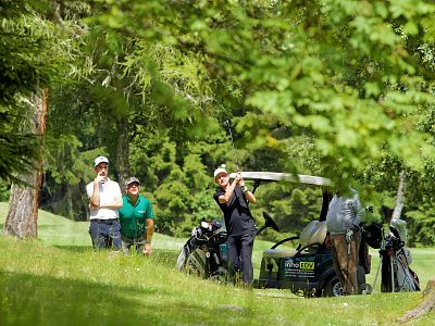 Golfers on a grassy course, surrounded by trees, with one in mid-swing and others observing near a golf cart.