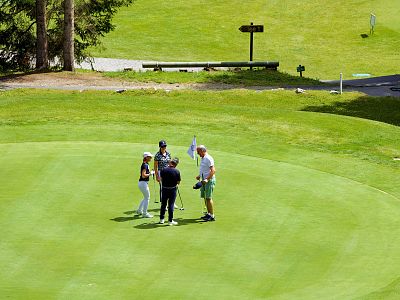 Four golfers stand together on a green, discussing strategy. The scene is surrounded by lush grass and nearby trees on a sunny day.