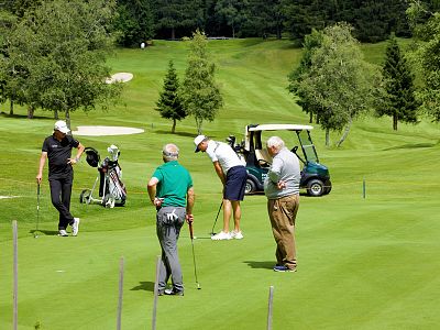 Four golfers stand on a green golf course with clubs. Two older and two younger men, with golf cart and trees in the background, are chatting.
