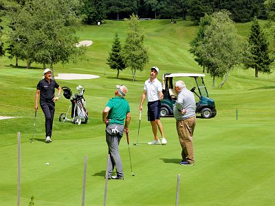 Four men on a golf course, standing on green grass, with trees and a golf cart in the background, enjoying a day of golf.