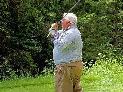 Elderly man in golf attire swinging a club on a green course, surrounded by dense trees and a grassy field, demonstrating a focused posture.