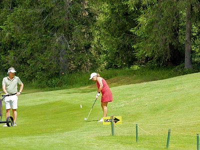 A woman in a red dress plays golf on a lush course, swinging her club. A man stands nearby with a golf bag, watching the action.