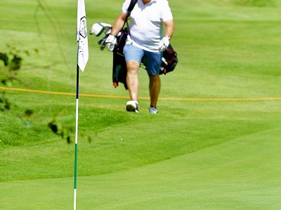 A man in casual attire walks on a lush golf course, carrying golf bags. A flagstick stands in the foreground on the freshly manicured green.