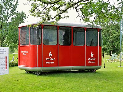A vintage red cable car cabin with KitzSki logo is placed on grass, surrounded by trees. It has large windows and a side sign with information.