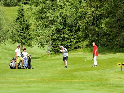 Four people on a vibrant green golf course; one prepares to swing, others watch. The scene is surrounded by trees under a clear sky.