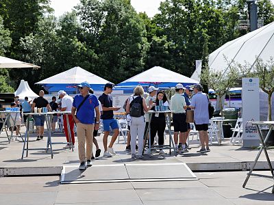 People at an outdoor event with tables and umbrellas, enjoying a sunny day. Trees and a clear sky form the backdrop of this casual social scene.