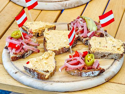 Slices of bread with spreads on a wooden platter, garnished with Austrian flags, pickled onions, peppers, and cheese, presented on a wooden table.