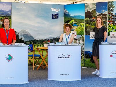 Three women stand behind booths at a trade fair, with landscape posters behind them. They appear friendly and are in a professional setting.