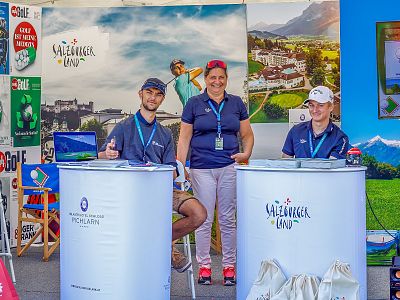 Three individuals at a booth with "Salzburger Land" branding, smiling and posing. Various promotional materials and a scenic backdrop are visible.