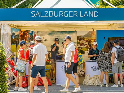 People gather at a 'Salzburger Land' booth showcasing scenic images and offering brochures, promoting tourism at an outdoor event.