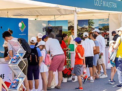 Mehrere Menschen versammeln sich an einem Messestand im Freien. Der Stand ist mit Schildern für Golf in Österreich dekoriert. Einige tragen Freizeitkleidung.