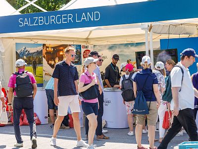 People at an outdoor event with a booth promoting Salzburg Land, featuring brochures and scenic images under a sunny blue sky.