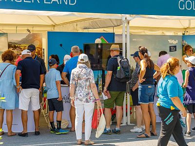 People gathered at an outdoor booth under a canopy, viewing displays. The event seems related to golf, with signage and people showing interest in activities.