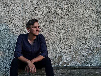 Ein Mann sitzt nachdenklich vor einer rauen, grauen Wand. Er trägt ein dunkles Hemd und schwarze Hosen, seine Hände liegen locker auf seinen Knien.
