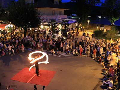 Nighttime outdoor scene with a fire performer in front of a seated crowd, using flames to create trails of light. Spectators are sitting and standing nearby.