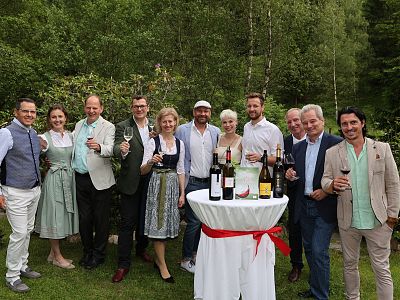 A group of people standing outdoors on a lawn, smiling and holding wine glasses. A table with wine bottles is in the center. Trees and greenery in the background.