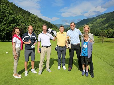 A group of people smiling on a golf course, standing with trees and mountains in the background, holding golf clubs and a trophy.