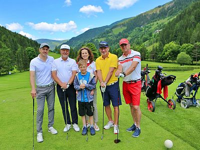 A group of six people, including a child, stand on a golf course with clubs, surrounded by trees and hills under a blue sky with clouds.