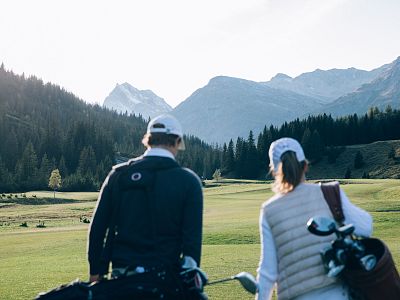 Ein Mann und eine Frau tragen Golfschläger über eine grüne Wiese. Im Hintergrund sind bewaldete Hügel und hohe Berge unter klarem Himmel zu sehen.