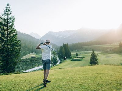 Ein Mann schwingt auf einem sonnigen Golfplatz seinen Golfschläger, umgeben von grünen Wiesen und hohen Bergen im Hintergrund, unter klarem, blauem Himmel.