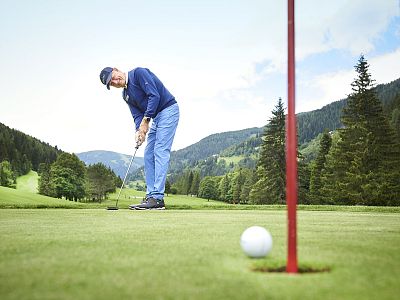 A golfer in blue attire putts on a green course, with mountains and trees in the background under a blue sky.