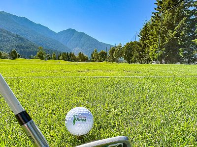 A golf ball on a lush green golf course with clubs nearby, against a backdrop of mountains and a clear blue sky. Trees line the fairway, enhancing the view.