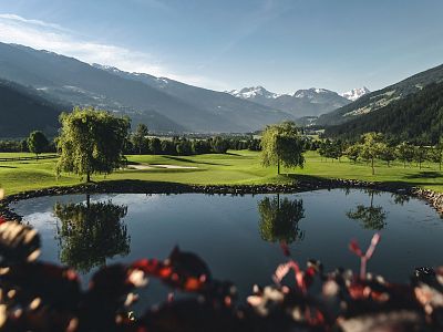 A golf course with a clear pond is surrounded by mountains and green fields under a bright blue sky, creating a peaceful and scenic landscape.