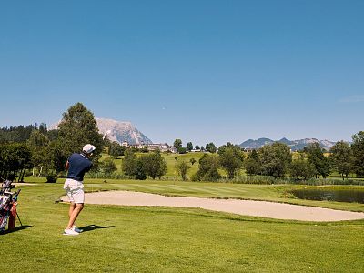 A golfer swings on a green course with mountains and blue sky in the background, embodying a peaceful outdoor sporting scene.