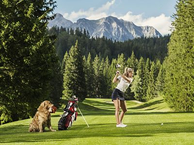 Woman golfing on a green course with a dog sitting beside her. Mountains and tall trees are in the background under a clear sky.