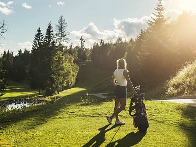 A woman on a golf course, standing with a cart against a backdrop of trees and grass, under a setting sun creating a warm and serene atmosphere.