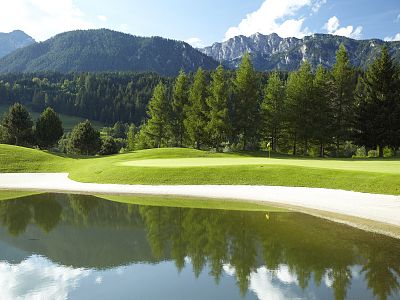 A serene golf course with lush greenery, sand trap, and a reflecting pond. Surrounding mountains and pine trees under a bright blue sky create a picturesque scene.