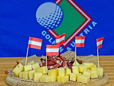 A platter with cheese cubes and ham slices, adorned with small Austrian flags, in front of a 'Golf in Austria' flag on a wooden table.
