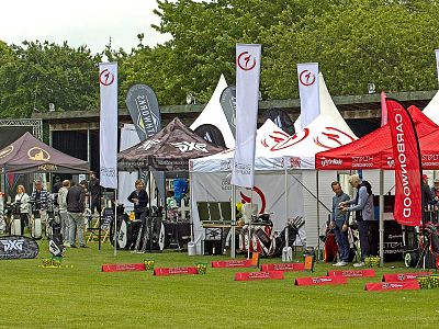 Outdoor expo with branded tents displaying golf gear and equipment. People are visiting various stalls, surrounded by lush greenery and clear skies.