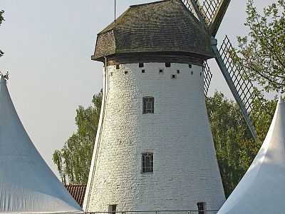 White cylindrical windmill with dark roof and sails, surrounded by large white tents and trees under a clear sky.